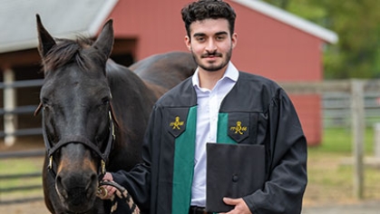 A graduate in regalia standing with a horse.